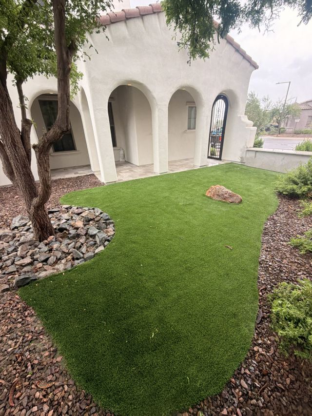 Curved artificial turf front yard edged with rock, with a boulder and a ring of river stone around a tree.