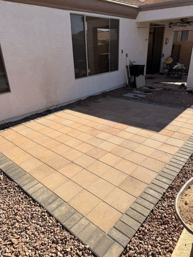 Tan square paver patio framed by a row of dark gray border pavers, set in rock beside a stucco home.