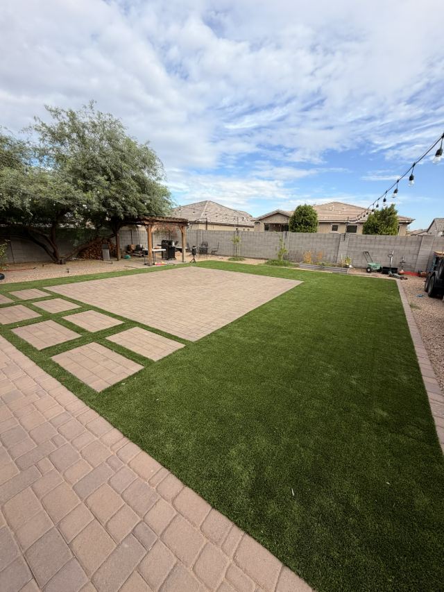 Large tan paver patio with a row of rectangular paver stepping pads set into artificial turf.